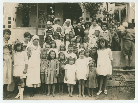 Photographie d'un «premier groupe d'enfants orphelins après les turbulences de la guerre», encadrés par des infirmières et autres adultes – avec légende manuscrite et tampons de la Croix Blanche polonaise au verso